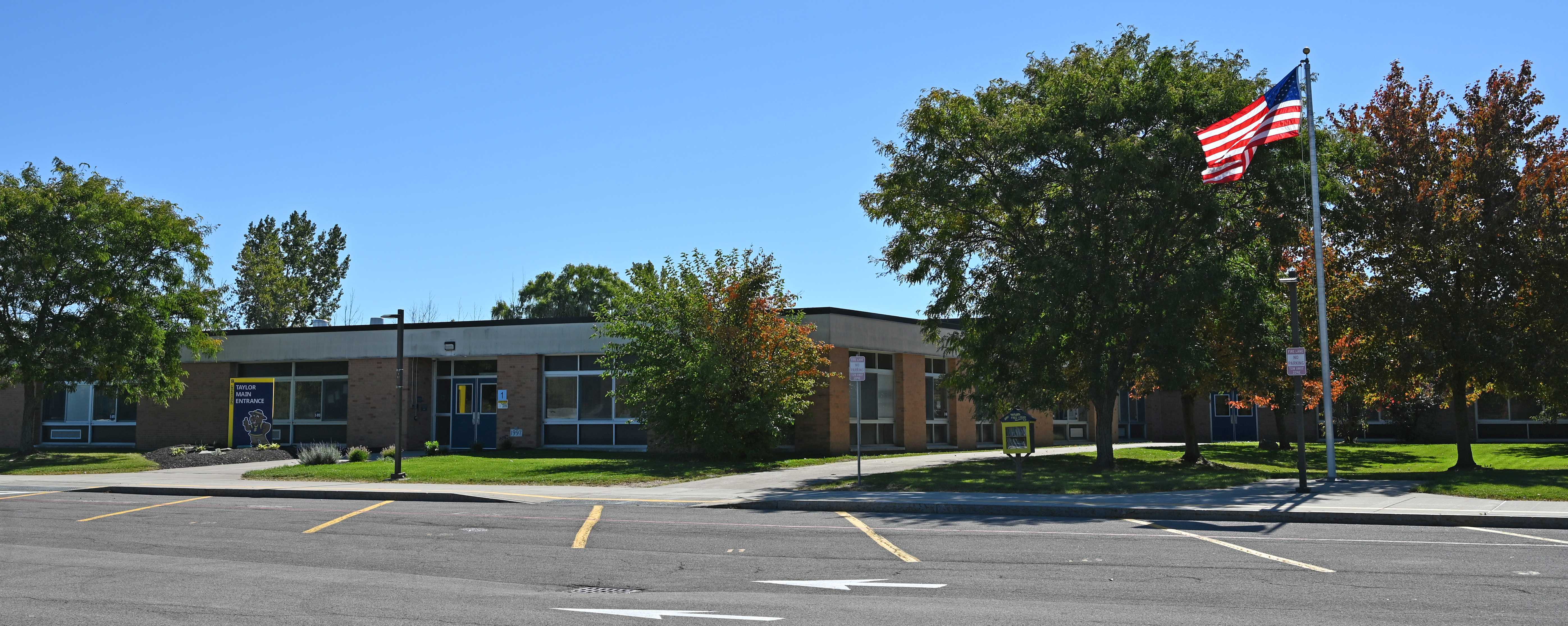 Taylor Elementary under a clear blue sky, surrounded by green trees and an empty parking lot.